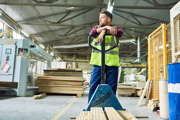 Full length portrait of young man wearing reflective jacket leaning on cart while moving wooden boards  in factory warehouse, copy space