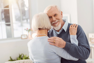 Happy husband. Joyful elderly man smiling at the camera while dancing with his beloved wife, hugging her tightly