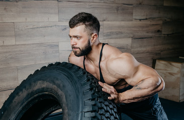 Strong muscular shredded man with tattoos and beard pushing a tire in a black tank top and grey...
