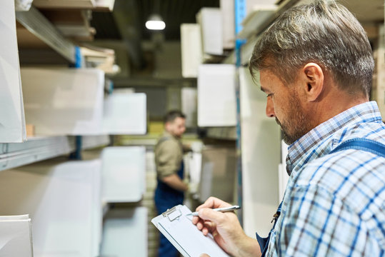 Portrait of mature worker  holding clipboard making list of materials standing in storage room with another man in background, copy space