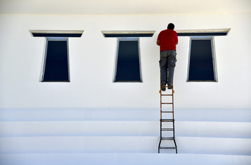 A man perched on a ladder is painting white a wall of a building