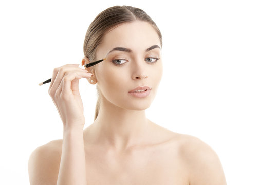 Studio Shot Of A Beautiful Young Woman Applying Makeup To Her Eyebrow While Standing At Isolated Background.