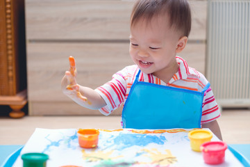 Cute smiling little Asian 18 months / 1 year old toddler baby boy child finger painting with hands and watercolors, kid painting at home, Creative play for toddlers, Montessori education concept