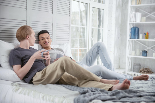 Life together. Positive energetic gay couple lying on bed while staring at each other and holding cups