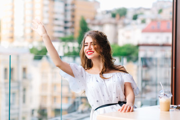 Fototapeta premium Beautiful girl with long hair is sittting at table on the terrace in cafe. She wears a white dress with naked shoulders and red lipstick . She is calling for waiter.