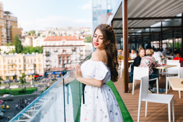 Beautiful girl with long hair stands  on the terrace in cafe . She wears a white dress with bare shoulders and red lipstick . She has ba light smile and looks down.