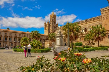 Historical Palermo Cathedral with hugging couple ot tourists the view thru beautiful mediterranean flowers with palm and beautiful sunny sky with clouds - Italy Sicily Aincient Europe 