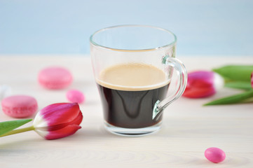 Transparent cup with coffee surrounded by pink tulips and pink macaroons. Light background. Close-up. Macro photography.