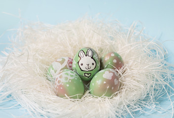 Colorful Easter eggs handpainted with floral decoration and a bunny, on a white nest. Pastel blue background. Perfect for your festive greetings cards.