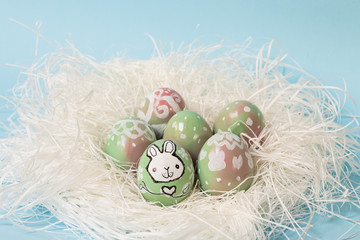 Colorful Easter eggs handpainted with floral decoration and a bunny, on a white nest. Pastel blue background. Perfect for your festive greetings cards.