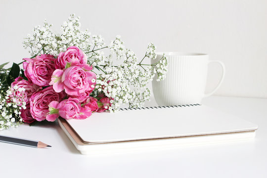 Styled stock photo. Closeup of wedding bouquet made of pink roses and baby's breath, Gypsophila flowers lying on white table. Feminine still life, blank notebook, pencil and white cup of coffee.