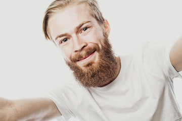 I love selfie! bearded man holding smartphone and making selfie and smiling while standing against white background.