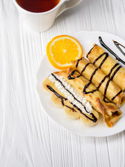 Pancakes with banana, whipped cream decorated with chocolate syrup on white wooden background and cup of tea. Top view.