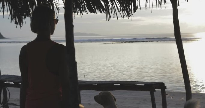young joyful dreaming woman or girl (silhouette) sitting on the beach or coast under conopy (awning, sunshade), looking at sunset or horizon, sea or ocean (water), beautiful sky, wearing sunglasses