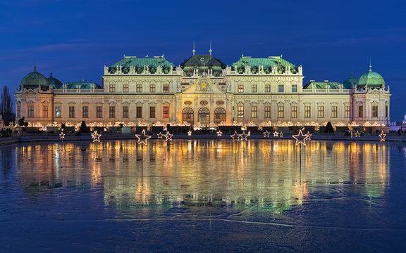 Upper Belvedere Palace with Christmas Village reflecting in the pond covered with wet ice in twilight, Vienna, Austria