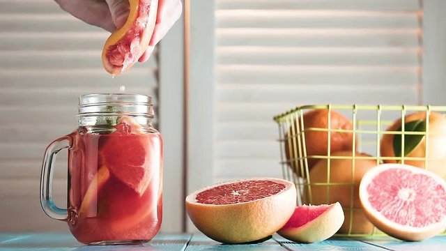 Man's hand squeezes juice from grapefruit into mason jar, slow motion. Fresh grapefruits, cocktail with grapefruit slices and mint on rustic blue wooden table opposite the blinds.