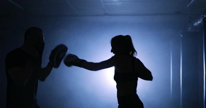 Silhouettes of a female boxer punching a boxing bag with boxing gloves in a smoky gym