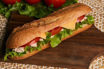 Fresh submarine sandwich with chicken fillet, cheese, fresh tomatoes, lettuce, cucumbers and onions on wooden background