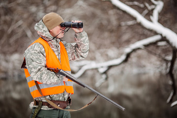 Male hunter in camouflage, armed with a rifle, standing in a snowy winter forest with duck prey