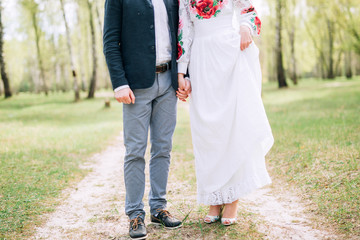 Fototapeta premium the groom in a blue suit holds a bride in the hand in a white embroidered dress with a veil on a meadow