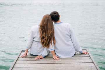 close-up of a girl and a guy in jeans and white embroidered shirts sitting on a wooden bridge and looking at the lake