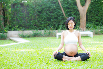 Beautiful pregnant asian young woman relax in the park, girl with belly sitting with crossed leg on grass and yoga exercise for wellness, copy space.