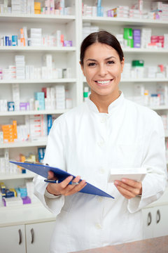 Medicine, pharmaceutics, health care and people concept - Laughing young pharmacist holding a clipboard and box of medications. Looking at camera