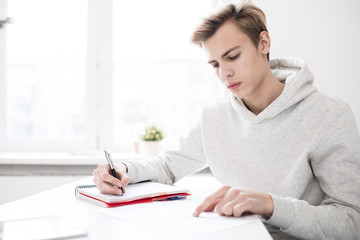 Handsome Caucasian teenage boy in hoodie sitting at desk in library and doing assignment in his notepad