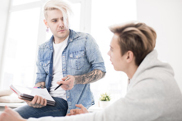 Fototapeta premium Tattooed adult student with dyed blond hair sitting on windowsill and discussing assignment with young male college mate