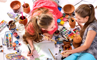 Authentic artist children girl paints with set palette watercolor paints palette and brush in morning sunlight. Painting in studio on floor. Older sister teaches younger to draw still lifes.