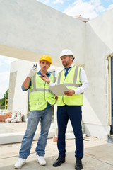 Full length portrait of foreman and supervisor discussing construction on site, both wearing...