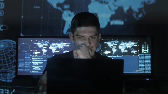 Hacker programmer in glasses is working on computer while blue binary code characters reflect on his face in cyber security center filled with display screens.