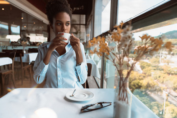 Pensive exquisite African American girl holding the cup of coffee while sitting at the table of a luxury restaurant on the top-floor; young thoughtful black female with the cup of cocoa in a cafe