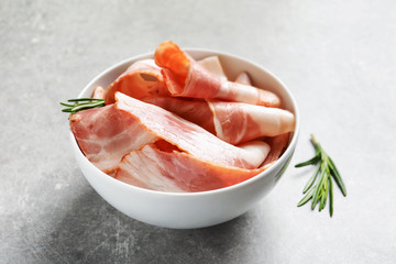Bowl with raw bacon rashers on grey background