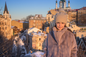 Fototapeta premium beautiful girl stands in a gray fur coat and a knitted beige hat on the background of houses