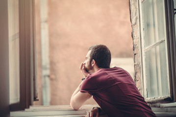 Melancholic and lethargic man looking outside through a big window, contemplating his emotional or...