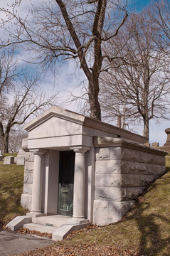 A mausoleum in a cemetery on a bright winter day. Blue skies with white clouds can be seen through the bare trees