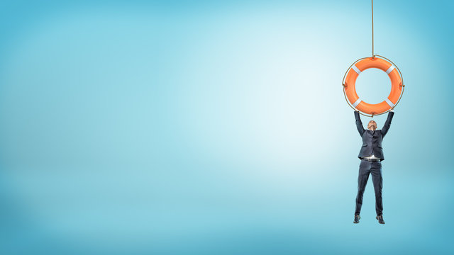 A tiny businessman holds on to a giant orange life buoy with both arms on a blue background.