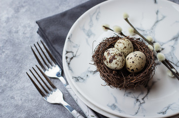 Festive table setting for holiday Easter dinner on gray stone concrete table with copyspace. Quail eggs and willow on plate