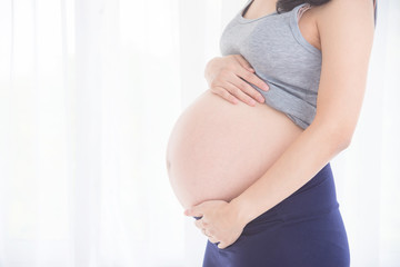 Cropped image of asian pregnant woman,holding her belly in front of bright window.