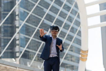 Young Asian businessman in a suit wear and virtual reality glasses who trying to touch something with his hand with city town background. Concept Technology and Simulation Business