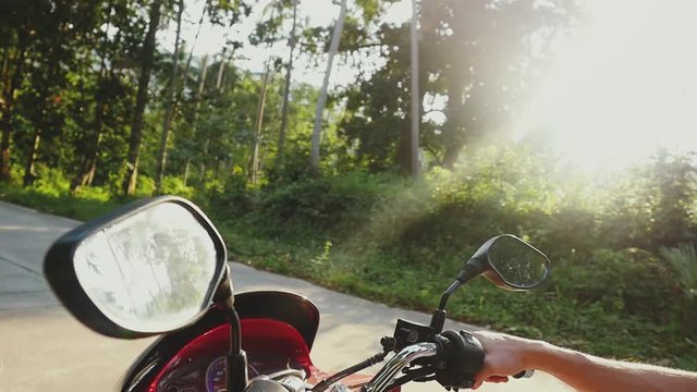 Young handsome man in sunglasses driving motorbike on tropical sunny road through the sun with beautiful lens flare effects in a jungle. slow motion. Close up. 1920x1080