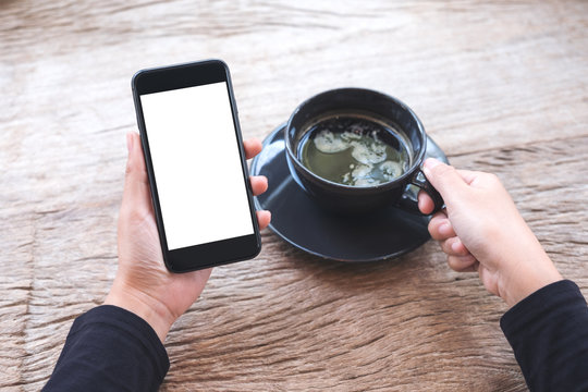 Mockup image of hands holding black mobile phone with blank white desktop screen and a cup of coffee on wooden table in cafe