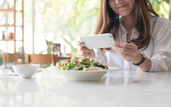 Closeup image of an asian woman using smartphone to taking photo of salad in a white plate on table in cafe
