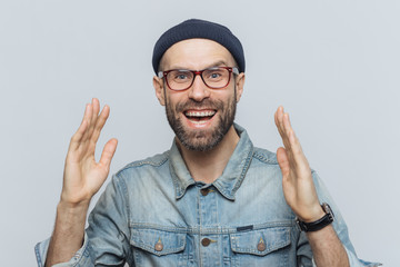 Handsome happy man raises hands with excitment, has overjoyed expression, looks with delighted face, has thick beard and mustache. Glad man in eyewear poses against white background, has fun