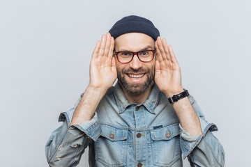 Portrait of happy hipster guy with thick beard and mustache wears stylish hat and glasses, keeps hands near face, tries to see something in darkness, isolated over grey background, has fun indoor
