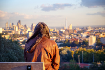 Back view of a female tourist looking over London city skyline from Parliament Hill in Hampstead Heath at sunset