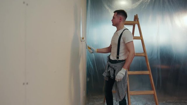 Attractive young man in a casual outfit in the process of wall painting. He uses a professional tools. Apartment in the process of refit. Lifehack, western-style renovation. Slow motion, camera