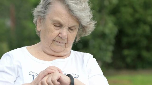 Black wristband. Mature elderly woman dressed in white t-shirt looks at the results of physical activity using a wristband fitness tracker. Positive person. Excellent mood