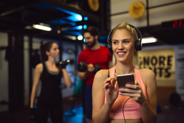 Plakat Sportswoman with smartphone listening to music in gym. Friends are talking behind, blurred in background.